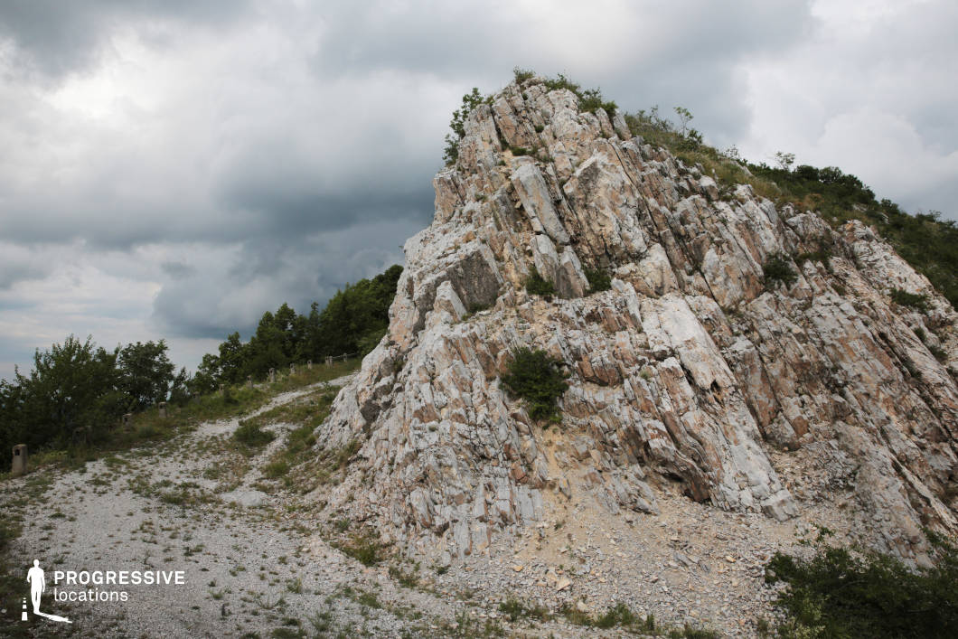 This dramatic rocky formation rises sharply with steep, angular striations and layered mineral textures, creating a visually striking natural structure against a backdrop of rugged terrain. The sparse vegetation clinging to the rock face and the gravel path curving around its base add to the raw, untouched quality of the setting. Ideal for survival films, adventure stories, or fantasy epics, this location suits scenes of perilous journeys, hidden mountain trails, duels on elevated ground, or solitary moments of challenge and triumph.