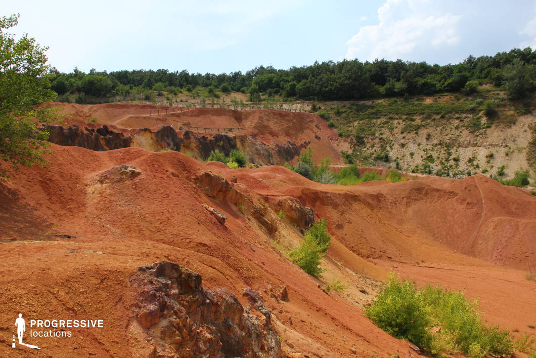 This striking location showcases a vibrant terrain of red and ochre hills, eroded gullies, and exposed rock formations within a former open-pit mine. The bold coloration and rugged texture create a dramatic, alien-like atmosphere, contrasting sharply with the surrounding green vegetation. Its unique visual character makes it ideal for fantasy, sci-fi, dystopian, or western-style productions, especially scenes set in desolate or otherworldly environments, forgotten lands, or hostile terrains.