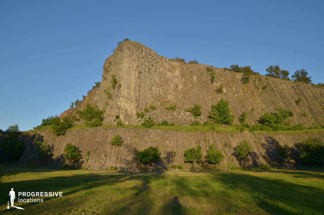 This dramatic rock formation features a towering basalt cliff with distinct vertical columns, rising from a tiered slope that adds natural texture and depth. The precise, geometric striations of the rock create a sculptural, almost otherworldly backdrop set against an open, grassy foreground. The bold geological features and elevated perspective make this a powerful filming location for adventure, fantasy, or post-apocalyptic genres—ideal for establishing shots, confrontational standoffs, sacred sites, or moments of revelation.