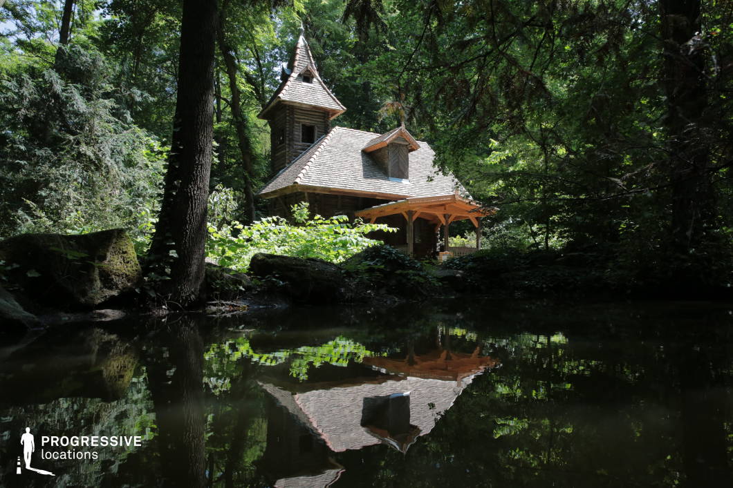 This enchanting forest location features a small, towered wooden cottage nestled among dense trees and reflected in a still, shadowed pond in the foreground. The structure is built with natural timber and topped with a steep, shingled roof, blending harmoniously with the surrounding vegetation. The architecture includes a covered porch and pointed dormers, enhancing the storybook-like quality of the setting. Framed by overhanging branches and moss-covered stones, the composition emphasizes intimacy and seclusion. Ideal for fantasy, mystery, or children’s film productions, this location offers a richly atmospheric and immersive woodland backdrop for scenes requiring isolation or magical ambiance.