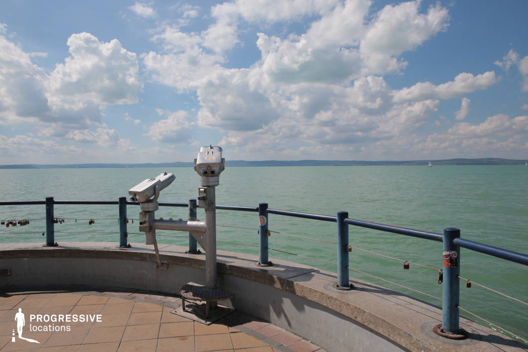 This location presents a curved observation point with mounted metal binoculars, enclosed by a simple blue railing set against a broad expanse of calm water. The tiled platform and clean lines of the structure suggest a contemporary lakeside promenade or scenic stop. The sweeping horizon and uncluttered composition make it a versatile backdrop for a variety of visual moods. Ideal for travel-related films, romance scenes, introspective character moments, or transitions involving journey and reflection, this lookout offers cinematic framing with both openness and clarity.