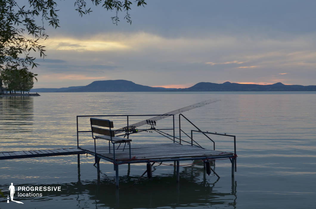 This serene lakeside setting features a narrow wooden pier extending over calm waters, framed by a mountainous horizon and fading light. The stillness of the lake, distant contours of flat-topped hills, and soft reflection create a tranquil and expansive composition. The scene’s natural symmetry and open sky make it ideal for introspective moments, romantic interludes, or meditative transitions in dramas, coming-of-age stories, or poetic travel films.