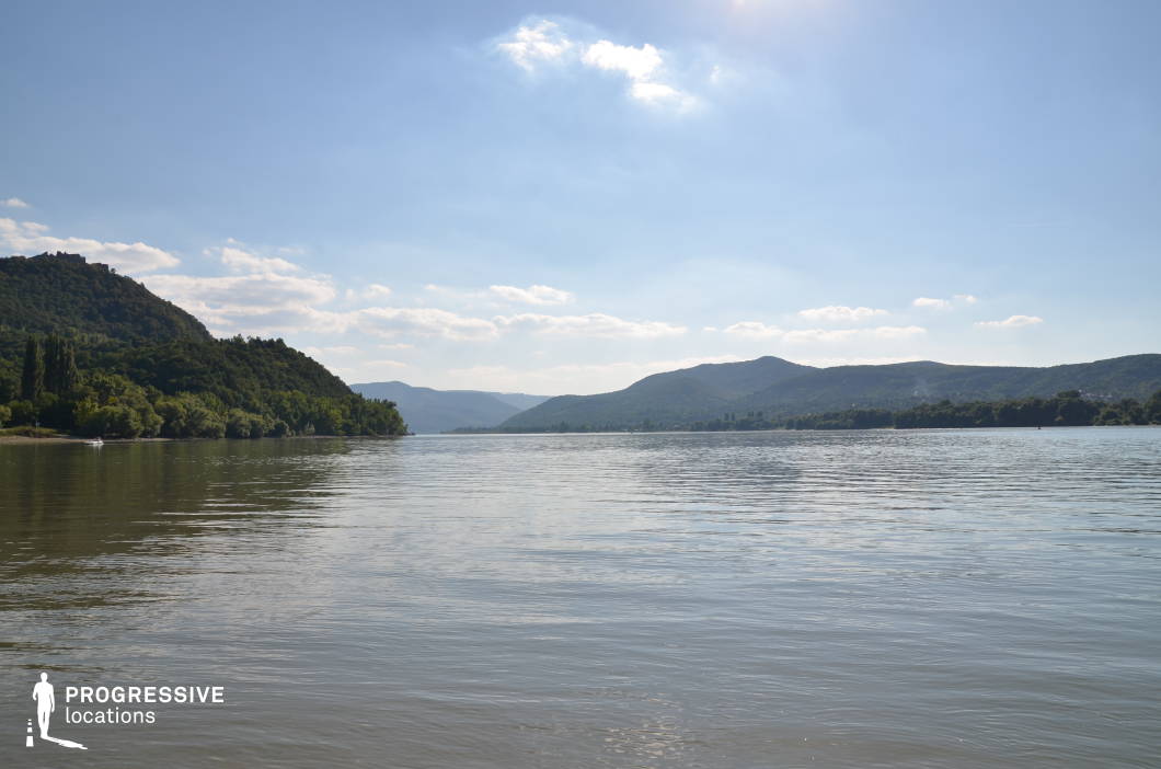 This panoramic riverscape captures a broad, tranquil waterway framed by rolling, forested hills on both sides, with a gently curving riverbank and a low island in the mid-distance. The natural symmetry, open horizon, and layered terrain create a cinematic depth that evokes calm, grandeur, or introspection. This location is ideal for epic journey scenes, river crossings, or reflective moments in period pieces, historical adventures, or nature-centered dramas.