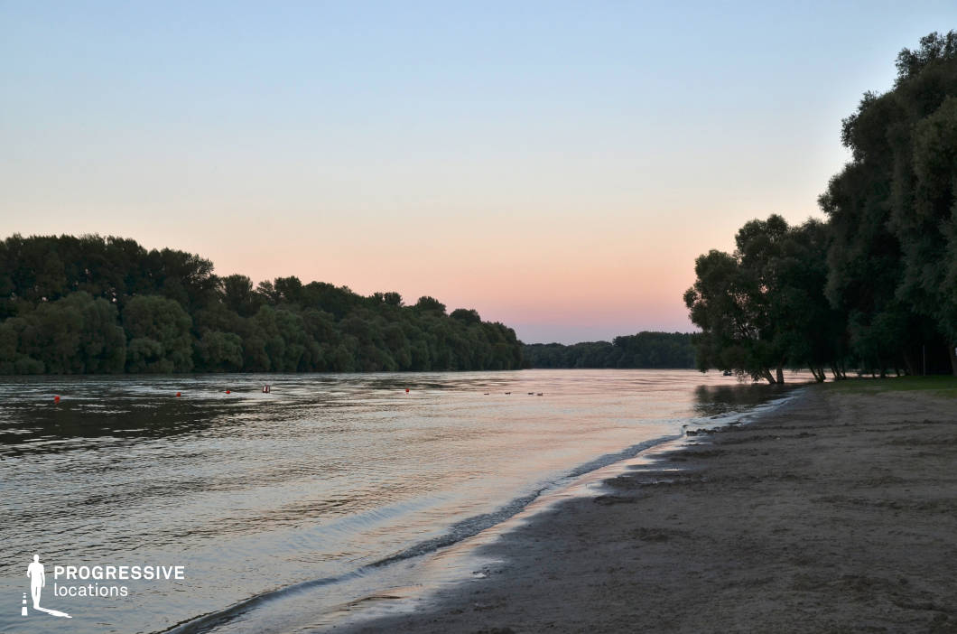This peaceful riverside location features a gentle, sandy shoreline curving along a broad, calm river, with densely forested banks on either side. The horizon glows with subtle pink and blue hues, creating a soft and contemplative atmosphere. The expansive water surface and natural framing by trees enhance the sense of isolation and serenity. This setting is ideal for reflective scenes, romantic moments, or story transitions in dramas, nature-focused films, or coming-of-age stories where a quiet, unspoiled riverscape enhances emotional depth.