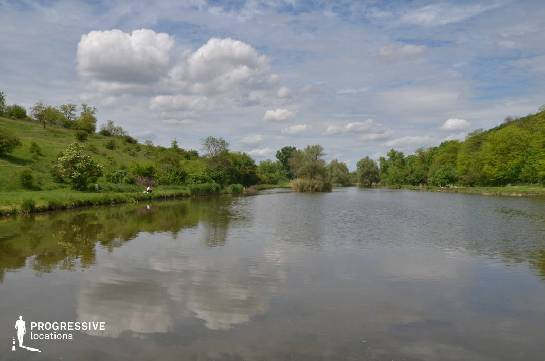 This peaceful countryside lake is framed by rolling grassy hills and densely vegetated shorelines, creating a harmonious blend of open space and natural enclosure. The still water reflects the surrounding greenery and gentle sky, while the irregular shoreline adds depth and visual interest. The landscape is characterized by soft contours and scattered trees, providing a pastoral backdrop with a serene, balanced composition. Ideal for family dramas, romantic scenes, or nature-themed productions, this location offers a calm, accessible setting with a timeless rural charm that supports both narrative and visual storytelling.
