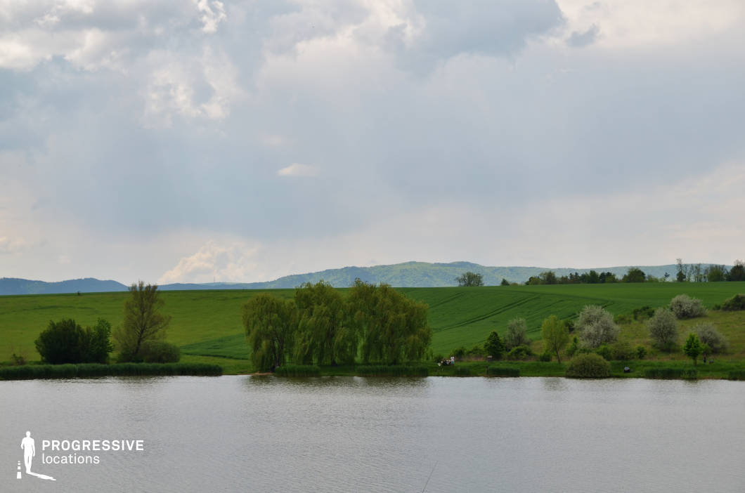 This location features a calm, reflective body of water bordered by lush, expansive fields that stretch into gentle hills in the background. The layered landscape, with a mix of open grassland and wooded patches, creates a serene and idyllic rural setting. Its open composition and natural symmetry make it ideal for filming peaceful interludes, contemplative moments, or scenes of escape and solitude. Perfect for romantic dramas, coming-of-age stories, or period pieces requiring a calm countryside atmosphere.
