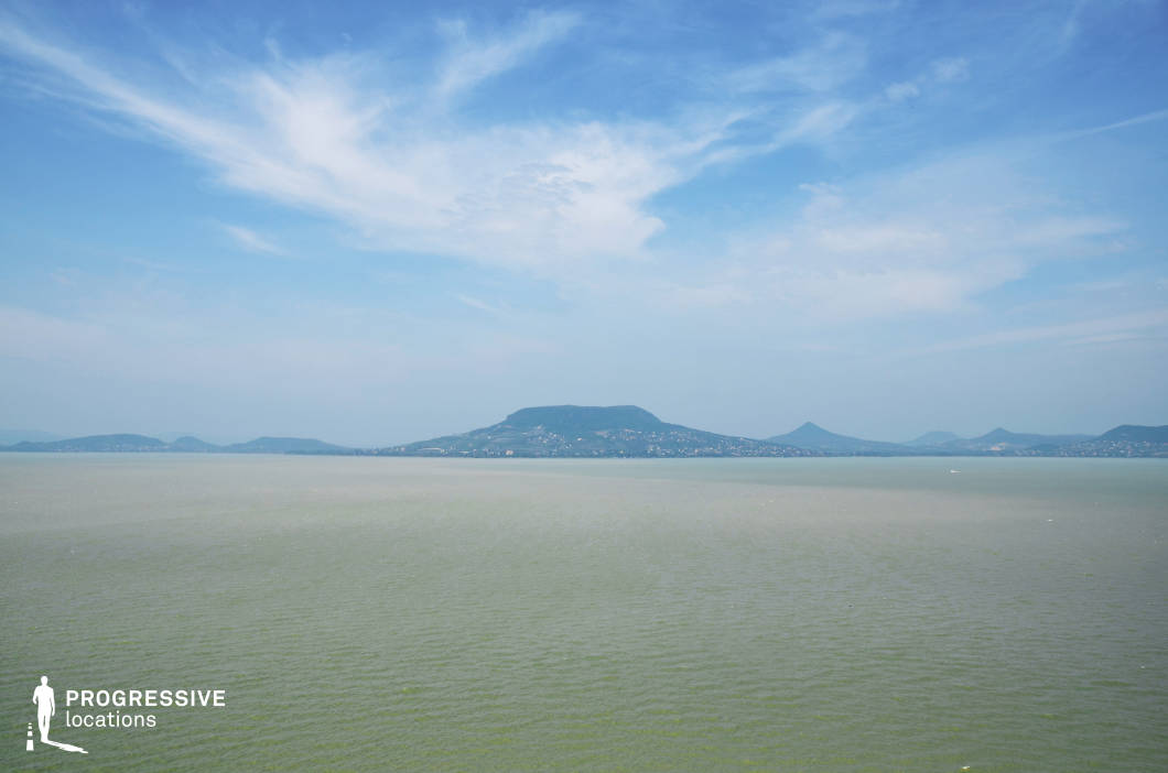 This expansive panoramic view features a calm, green-tinted lake stretching across the foreground, with a distinctive flat-topped hill and a chain of gently sloping elevations defining the distant horizon. The scene is characterized by its layered depth and horizontal visual rhythm, with subtle tonal gradients across the water enhancing the composition's sense of scale and openness. The landscape is free from built structures in the immediate foreground, offering an uninterrupted natural tableau. This location is ideal for wide establishing shots, romantic or contemplative scenes, and any film genres requiring tranquil natural vistas, such as dramas, road movies, or nature-centric productions.