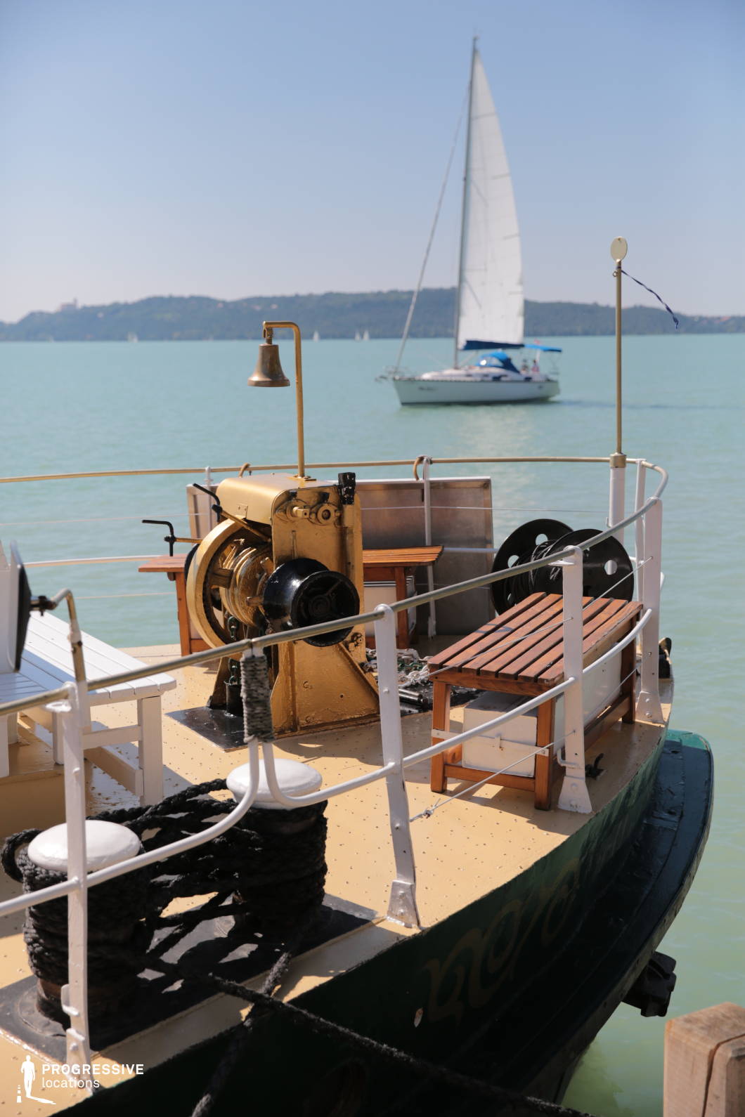 This image captures the stern of a vintage vessel with brass fittings, rope cleats, wooden benches, and a mechanical winch, all enclosed by a classic white railing. The calm, pale turquoise water stretches into the distance, with a sailboat and a low wooded shoreline providing a scenic backdrop. The composition blends nautical nostalgia with tranquility. This location is ideal for filming period maritime scenes, romantic interludes, reflective monologues, or travel montages, especially in productions set on inland waters or evoking a timeless lakeside charm.