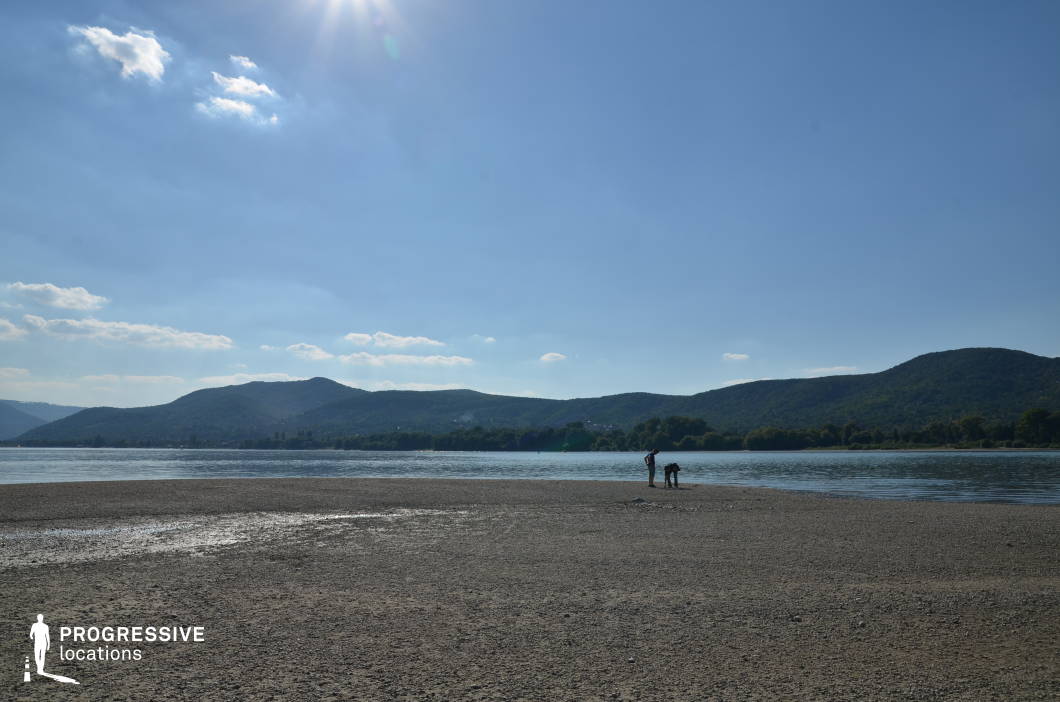 This wide, open riverbank stretches into the distance with a gentle curve of water bordered by forested hills and a vast, pebbled shore. The low perspective emphasizes the natural scale and isolation of the area, while the surrounding landscape offers a sense of openness and transition. This location is ideal for scenes requiring emotional reflection, journeys, or pivotal character moments in dramas, nature-based narratives, or coming-of-age films, particularly those emphasizing solitude, escape, or the crossing of natural boundaries.