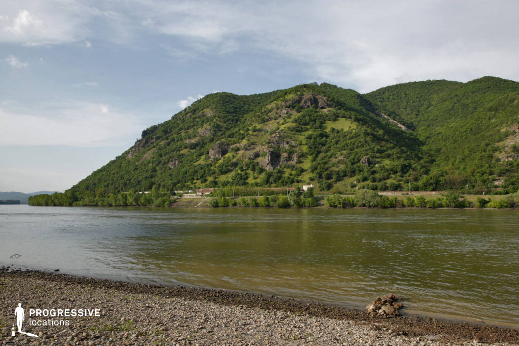This location presents a tranquil riverbank with a stony shoreline, looking across a broad, gently flowing river toward a lush, rugged hillside. The slopes are dotted with rocky outcrops and covered in dense vegetation, creating a striking contrast between water and elevated terrain. Ideal for nature-based storytelling, adventure, fantasy, or historical genres, this site can serve as the backdrop for river crossings, journeys, camp scenes, or serene establishing shots in untamed wilderness.