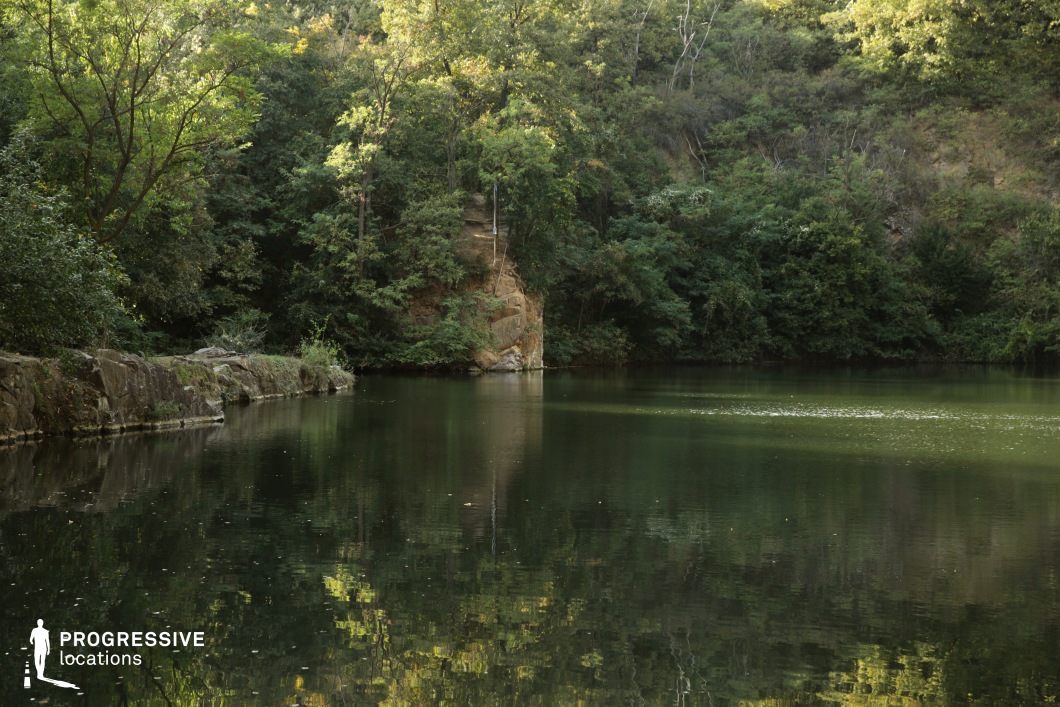 This intimate natural setting features a calm, reflective lake nestled within a dense forest, bordered by rocky outcrops and lush, varied vegetation. The water's surface captures a mirror-like reflection of the surrounding greenery, while the rocky shoreline adds textural contrast to the otherwise soft, organic forms. The terrain gently rises in the background, creating depth and a natural amphitheater effect that enhances acoustic and visual intimacy. This location is ideal for filming introspective, romantic, or mysterious scenes, particularly in drama or fantasy genres, where the sense of seclusion and natural harmony plays a central visual and atmospheric role.