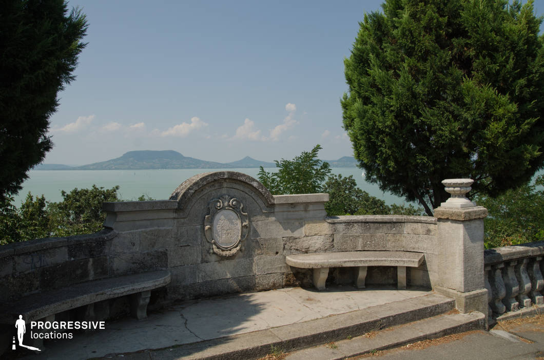 This elegant spot features a semicircular stone bench with carved details and a central crest, framed by classical balustrades and ornamental urns. Positioned at a scenic overlook, it opens to a calm, expansive body of water with distant hills on the horizon. The classical architectural elements lend a timeless and serene atmosphere, making this location ideal for romantic scenes, reflective moments, period dramas, or travel sequences. Its aesthetic balance and natural backdrop offer a graceful setting for emotionally charged or visually poetic shots.