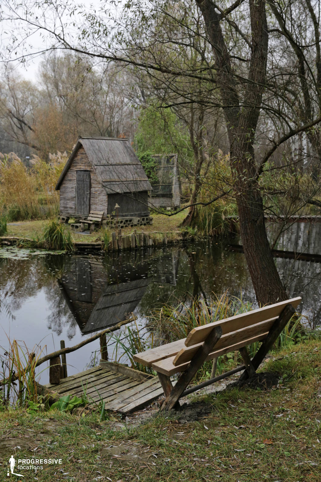 This peaceful lakeside spot features a weathered wooden hut set on the opposite shore, mirrored in the still water, with a small wooden dock and bench in the foreground. Surrounded by reeds, trees, and soft undergrowth, the location exudes calmness and seclusion. Perfect for introspective, romantic, or mysterious scenes, this setting works well for dramas, coming-of-age films, or thrillers involving hidden retreats, solitary moments, or quiet countryside life.