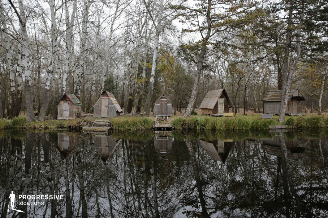 This secluded woodland location features a row of small, handmade wooden huts along the shore of a still, reflective lake, backed by dense trees with striking white trunks. The mirrored water surface and minimal modern interference lend the area an untouched, almost otherworldly quality. The rustic construction and natural setting evoke simplicity, isolation, or timelessness, making it ideal for filming scenes involving hermits, forgotten communities, survival stories, or magical realism in fantasy, folk horror, or arthouse productions.