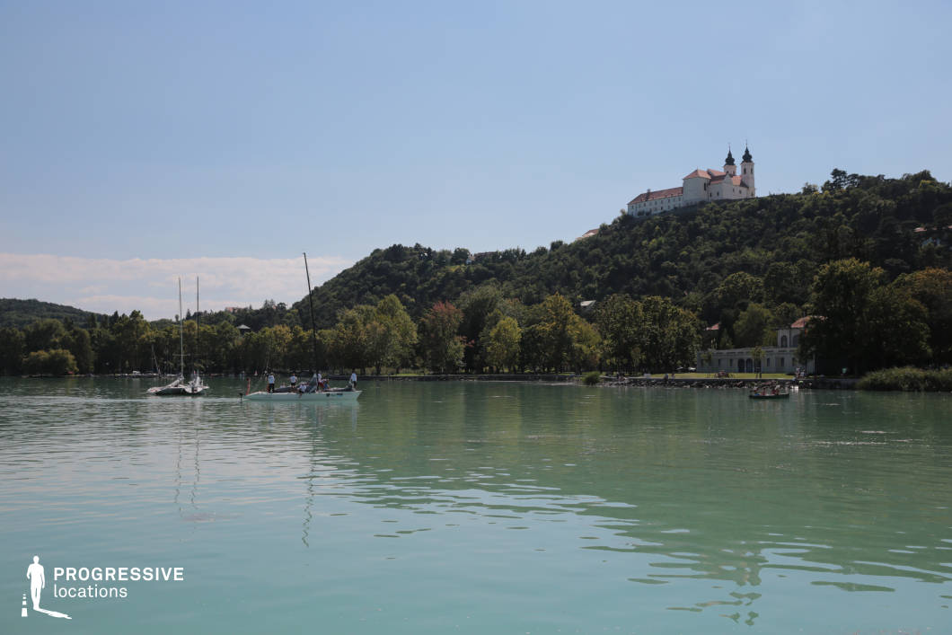 This scenic location presents a tranquil lakeshore with still, aquamarine waters set against a densely wooded hillside crowned by a prominent hilltop abbey. The architecture atop the hill provides a striking focal point, offering visual depth and a sense of historical or cultural significance. The gentle tree line along the shore adds balance and calm to the composition. This setting is perfect for romantic dramas, reflective character moments, or historical films, especially scenes involving arrival by water, peaceful transitions, or establishing shots that suggest spiritual or idyllic settings.