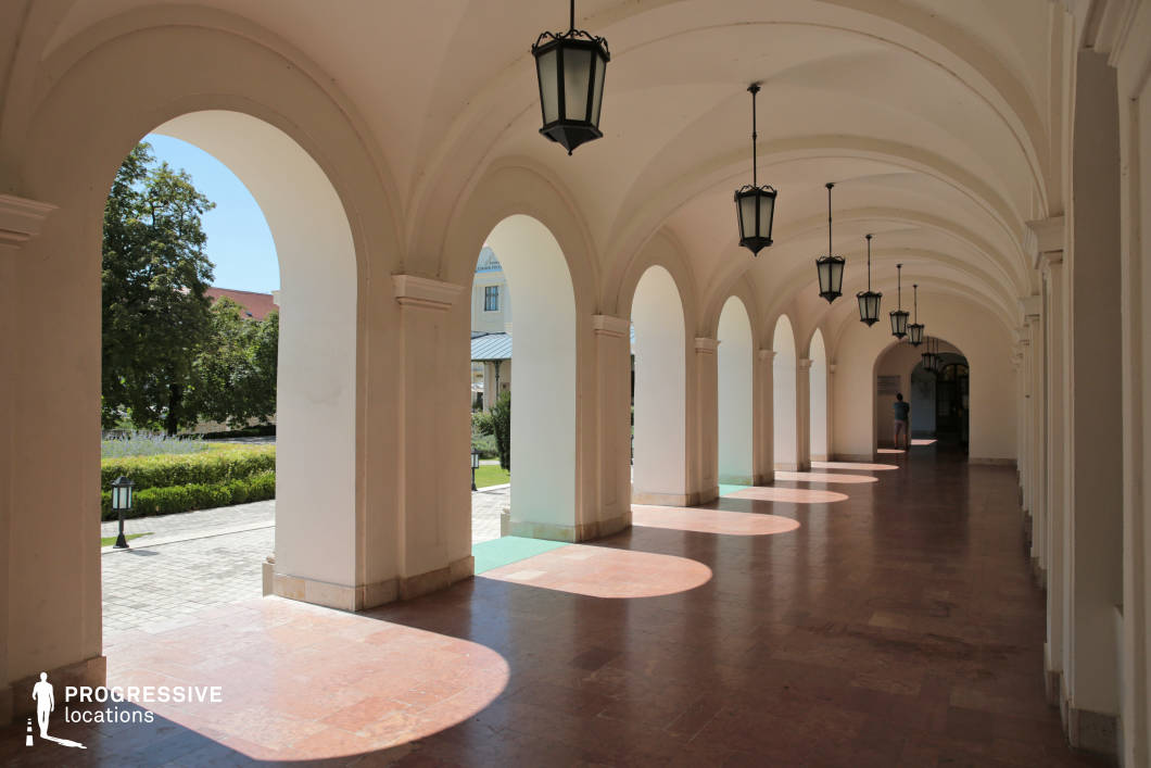 This covered arcade features a series of elegant, repeating arches supported by smooth, white columns, creating a symmetrical and structured architectural design. The vaulted ceiling is adorned with black wrought-iron lanterns hanging at regular intervals, adding a historical and refined aesthetic. The polished stone floor reflects the geometric patterns of the arches, enhancing the sense of depth and perspective. The open archways frame glimpses of a courtyard with manicured hedges and classic buildings in the background, contributing to a stately and European-style ambiance. This location is ideal for filming historical dramas, period pieces, or refined, atmospheric scenes in genres such as mystery, romance, or political thrillers, where a grand yet intimate setting is required.