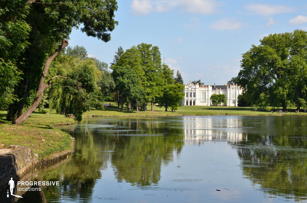 This serene landscape features a stately palace in the background, its white Neo-Gothic façade adorned with intricate architectural details and pointed roof elements. The palace is set within a vast, open park, with a tranquil lake in the foreground that reflects the surrounding scenery, creating a visually harmonious composition. The gently curving shoreline and well-maintained grounds provide a sense of openness and elegance. This location is ideal for filming romantic period pieces, fairy-tale-inspired stories, or introspective dramas, offering a dreamlike setting that blends aristocratic grandeur with natural beauty.
