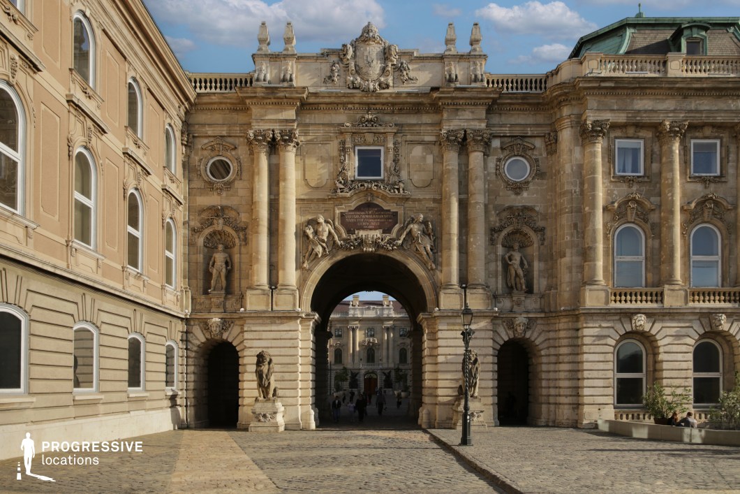 This grand Baroque-style gateway serves as a striking architectural centerpiece, featuring towering columns, intricately carved relief sculptures, and regal statues set within ornate niches. The imposing central archway, flanked by symmetrical decorative elements, creates a dramatic sense of depth, leading the eye toward the stately courtyard beyond. The weathered stone façade adds historical character, while the symmetrical arrangement of windows and sculpted figures enhances its formal elegance. The cobblestone foreground and classic street lamps contribute to the old-world atmosphere. This location is ideal for filming historical dramas, royal intrigue, and epic fantasy films, providing a regal and commanding backdrop suitable for grand entrances, confrontations, or ceremonial processions.