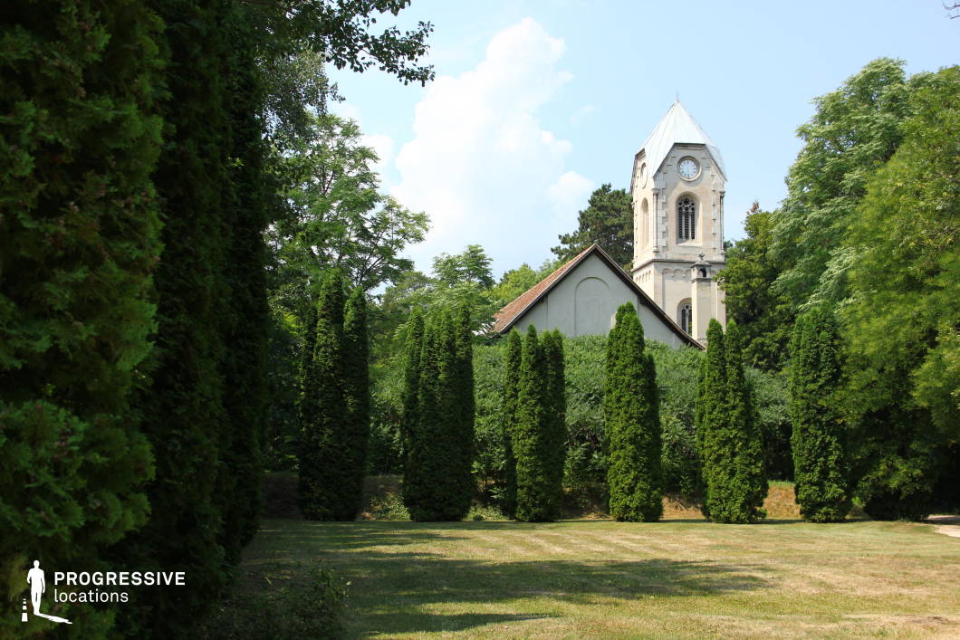 Nestled among tall, manicured evergreens, a historic chapel with a pointed clock tower emerges from the dense greenery. The light-colored stone facade of the tower, with arched windows and a steep metal roof, gives the building a serene yet imposing presence. The modest chapel structure, partially obscured by surrounding vegetation, features a simple pitched roof and smooth walls that contrast with the intricate details of the tower. The well-maintained grassy area in the foreground leads the eye toward the chapel, creating depth and a natural framing effect. This location is ideal for historical, mystery, or fantasy-themed films, offering a secluded and atmospheric backdrop for contemplative or dramatic scenes.