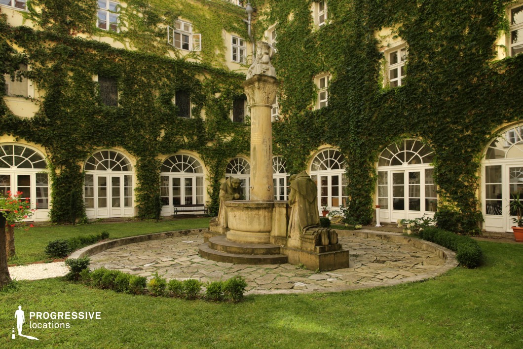 This image showcases a secluded courtyard enclosed by an elegant, ivy-clad building with arched windows and classic French doors. At the center stands a stone fountain adorned with sculpted figures, adding a sense of history and artistic detail. The surrounding greenery enhances the serene and intimate ambiance, while the well-maintained lawn and stone pathways contribute to the structured yet organic composition. The combination of classical architecture and lush natural elements makes this location ideal for filming historical dramas, romantic narratives, or period pieces, offering a refined and atmospheric setting perfect for elegant and contemplative scenes.