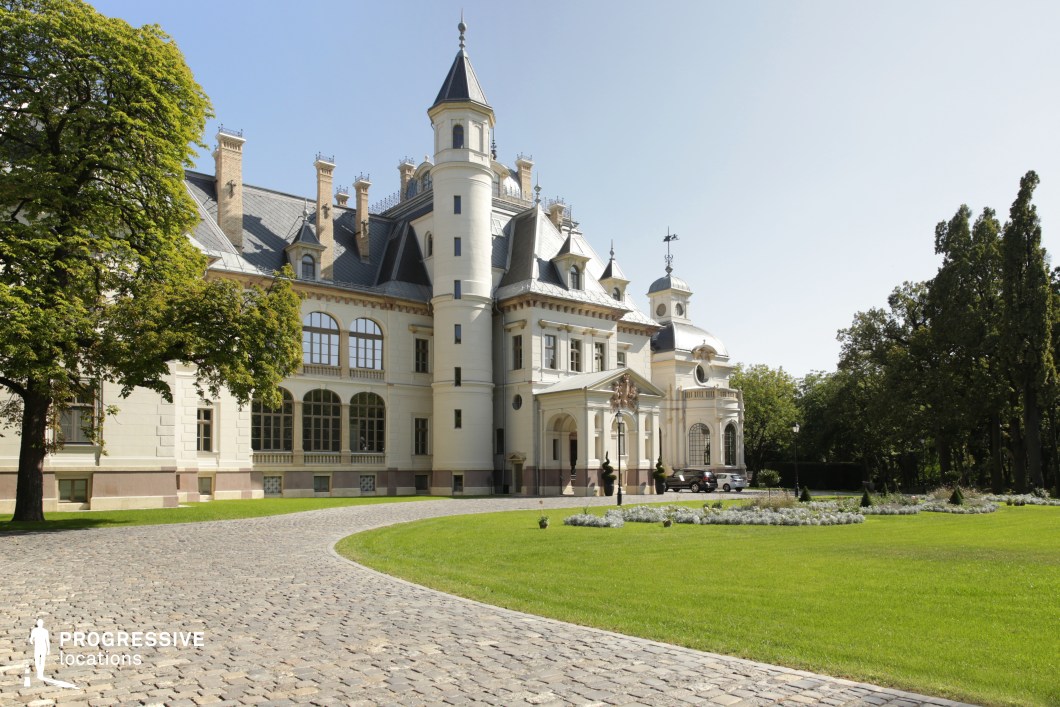 This image showcases an elegant château-style mansion with a distinctive blend of Gothic and Renaissance influences. The grand white facade is accentuated by a prominent cylindrical tower, steeply pitched roofs, decorative chimneys, and arched windows, exuding aristocratic charm. A gracefully curved cobblestone driveway leads to the main entrance, framed by a classical portico and intricate sculptural details. The surrounding landscaped garden, with neatly manicured lawns and ornamental plants, enhances the estate’s regal appeal. This location is ideal for filming period dramas, fantasy films, or historical romance stories, offering a stately and sophisticated backdrop for scenes of intrigue, luxury, or noble affairs.