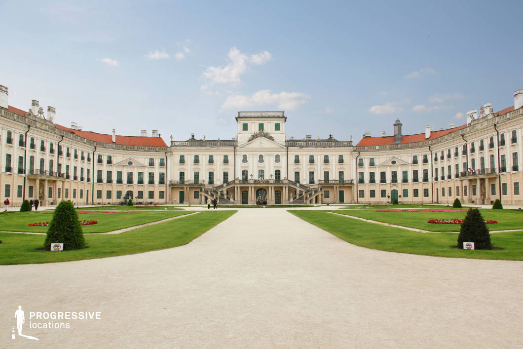 This image showcases a magnificent Baroque palace with an expansive, symmetrical courtyard framed by manicured gardens and a wide gravel pathway leading to the central building. The architectural composition features elegant arched windows, decorative balconies, and a grand double staircase ascending to the main entrance. The red-tiled roof contrasts with the pristine white facade, while ornamental sculptures and reliefs enhance the regal aesthetic. This location is ideal for filming period dramas, royal-themed productions, or historical epics, offering a stately and majestic backdrop for scenes of aristocratic life, formal gatherings, or political intrigue.