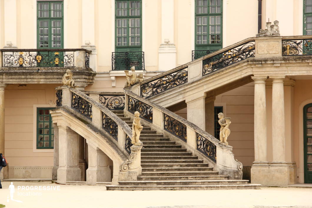 This image captures an elegant Baroque-style double staircase adorned with intricate wrought-iron railings featuring gilded embellishments. The stone balustrades are lined with classical statues, including cherubic figures and allegorical sculptures, adding to the grandeur of the design. The staircase leads to a terrace with arched openings and tall, green-framed windows, contributing to the palace’s aristocratic charm. The warm-toned facade, balanced by decorative columns and ornate moldings, reinforces the historical elegance of the setting. This location is ideal for filming period dramas, regal narratives, or historical romances, offering a stately backdrop for formal entrances, aristocratic gatherings, or moments of intrigue.