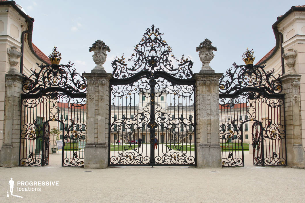 This image showcases an opulent wrought-iron gate adorned with intricate scrollwork, gilded accents, and crowned embellishments, serving as the grand entrance to a historic palace. The gate is framed by imposing stone pillars topped with sculpted urns, reinforcing the aristocratic grandeur of the setting. Beyond the gate, a symmetrical courtyard leads to a stately neoclassical palace with a pristine white facade, elegant arched windows, and a grand central staircase. The location is ideal for filming period dramas, historical epics, or regal narratives, offering a setting rich in prestige, exclusivity, and old-world sophistication.