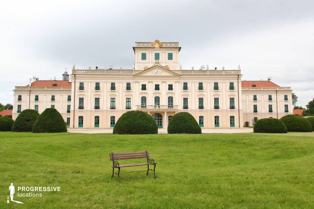 This image features a majestic Baroque palace with a symmetrical façade, elegant pastel tones, and ornate architectural detailing. The central clock and sculpted balustrades add a regal touch, while the evenly spaced windows and green shutters enhance the palace’s classical aesthetic. The foreground showcases a meticulously maintained lawn with geometrically shaped shrubs, creating a refined formal garden. The vast open space and balanced composition make this location ideal for filming historical dramas, aristocratic period pieces, or romantic films, providing a visually opulent and stately setting reminiscent of European royal residences.