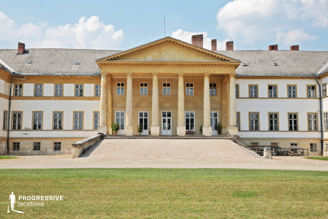 This image features a stately neoclassical palace with a symmetrical facade, dominated by a grand portico supported by six large, weathered columns. The building’s pale yellow and white exterior, along with its classical triangular pediment, exudes an air of faded aristocratic grandeur. Rows of evenly spaced windows frame the structure, with some showing signs of aging, adding to the historical character. A gently sloped gravel ramp leads up to the entrance, reinforcing the palace’s imposing presence. This location is ideal for filming historical dramas, political period pieces, or aristocratic narratives, offering a setting that conveys power, prestige, and old-world elegance.