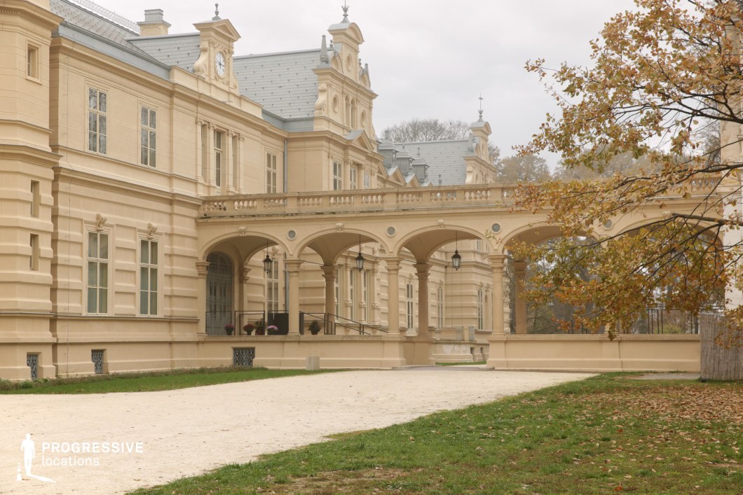 This image features a grand, meticulously restored palace with a neoclassical architectural style, characterized by its symmetrical facade, ornate detailing, and arched passageway supported by classical columns. The pale stone exterior is adorned with decorative moldings, sculpted reliefs, and a central clock, reinforcing the building’s aristocratic elegance. The balustraded terrace and hanging lanterns add refinement, while the soft natural setting enhances the stately atmosphere. This location is ideal for filming period dramas, historical romances, or royal narratives, offering a visually striking backdrop for scenes of opulence, diplomacy, or intrigue.