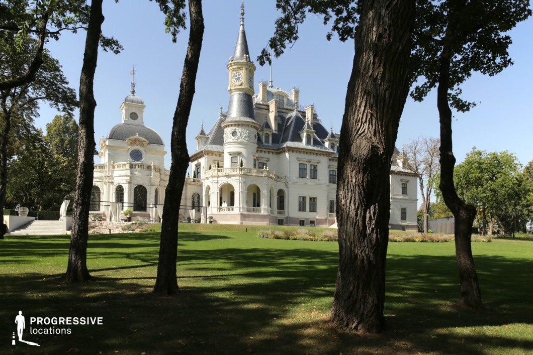 This image features a stunning château with an intricate blend of neo-Gothic and neo-Renaissance architectural elements. The grand white façade is adorned with decorative balconies, arched windows, and multiple turrets, including a prominent clock tower that enhances its regal presence. The surrounding parkland, framed by mature trees, provides a lush and serene setting, with the manicured lawn emphasizing the château’s elegance. The combination of refined architectural detailing and natural beauty makes this location ideal for filming period dramas, fantasy productions, or romantic narratives, offering a visually captivating and timeless setting.