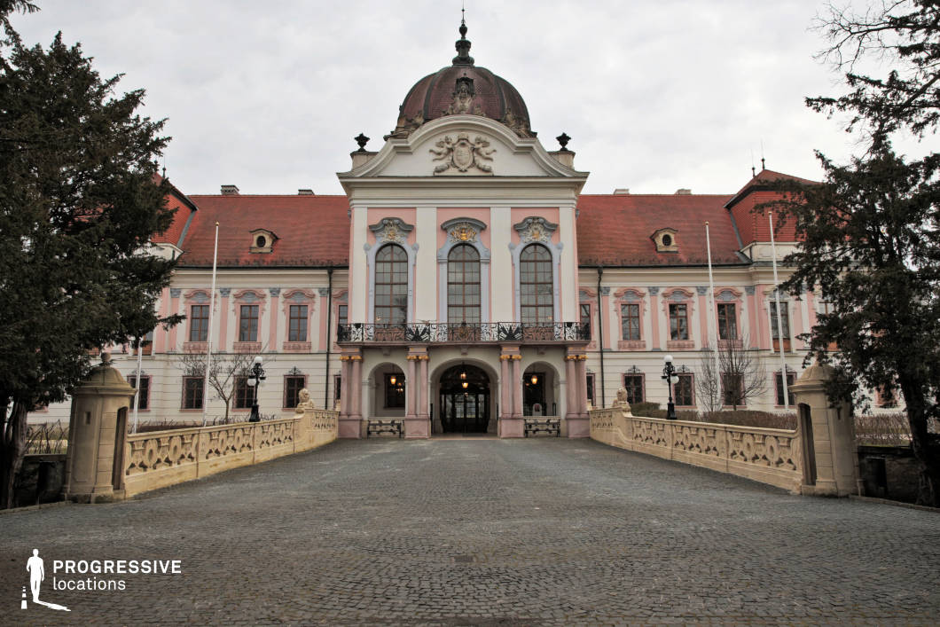 This image showcases an opulent Baroque palace with a striking symmetrical façade, adorned with intricate stucco decorations, tall arched windows, and a central dome. The soft pastel hues of the exterior, complemented by a red-tiled roof, enhance the regal elegance of the building. A cobblestone driveway leads to an ornate entrance framed by decorative stone balustrades and sculpted elements, reinforcing the palace’s aristocratic character. This location is ideal for filming period dramas, royal-themed productions, or historical narratives, offering a stately and visually grand setting that evokes the sophistication of European nobility.