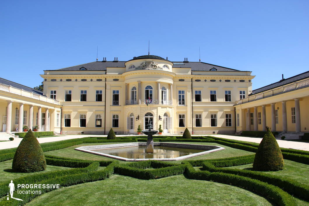 This image showcases a stately neoclassical mansion with a symmetrical design, featuring a curved central balcony, grand arched windows, and an ornate pediment adorned with sculptural reliefs. The facade is painted in a warm yellow hue, complemented by a dark mansard roof and classical columned walkways extending on either side. The foreground highlights a meticulously maintained French-style garden with geometric hedges, a decorative fountain, and cone-shaped topiaries, enhancing the estate’s aristocratic elegance. This location is ideal for filming period dramas, historical romances, or diplomatic settings, offering a refined and sophisticated backdrop for scenes of high society, political intrigue, or noble gatherings.