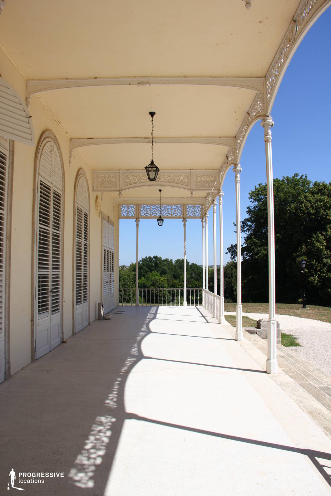 This image captures a beautifully crafted veranda attached to a historic mansion, featuring delicate ironwork and slender columns supporting an arched ceiling. The large, shuttered French doors and arched windows add a refined European charm, while the intricate patterns in the railings and decorative trim cast artistic shadows on the ground. The open-air structure offers a seamless connection to the surrounding landscape, creating a peaceful and sophisticated setting. This location is ideal for filming period dramas, romantic narratives, or introspective character-driven stories, providing a graceful and timeless atmosphere.