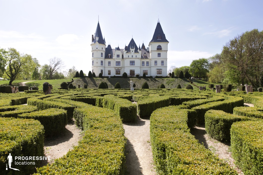 This image features a striking castle with a symmetrical design, steeply pitched roofs, and multiple conical towers, evoking a fairytale-like aesthetic. The white facade is accented with dark window frames and decorative elements, enhancing the castle’s elegant and timeless charm. In the foreground, a meticulously designed hedge maze with neatly trimmed pathways adds an element of mystery and grandeur. The elevated position of the castle, overlooking the maze, further enhances its commanding presence. This location is ideal for filming fantasy films, historical dramas, or adventure narratives, offering a visually captivating backdrop for royal intrigue, enchanted settings, or epic quests.