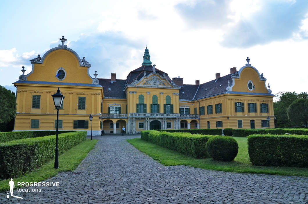 This grand baroque-style palace features a symmetrical yellow facade with ornate white detailing, arched windows, and decorative sculptural elements adorning the rooftop. The central section is accentuated by a pediment with intricate relief work and a green copper spire, adding a regal presence. The well-maintained garden, consisting of precisely trimmed hedges and a cobblestone pathway leading to the entrance, enhances the palace’s elegance. Traditional street lamps line the walkway, reinforcing the historical atmosphere. This location is ideal for period dramas, historical films, and aristocratic settings, offering an authentic European estate backdrop suitable for regal, romantic, or mystery-themed productions.