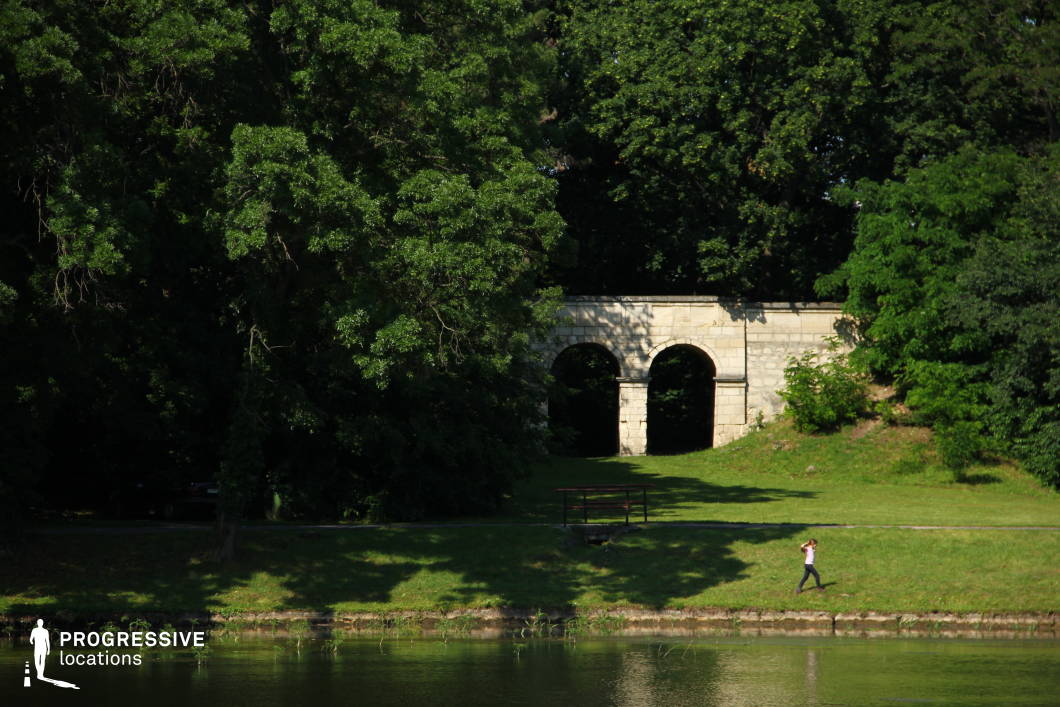 A tranquil park setting features a historic stone structure with two arched openings, partially concealed by lush greenery. The archway, built from weathered stone blocks, blends harmoniously into the natural landscape. A gently sloping grassy bank leads down to a calm lake, with a narrow stone edge marking the water’s boundary. The dense tree cover casts dappled shadows, adding depth and contrast to the scene. This location is ideal for historical dramas, mystery films, or contemplative, atmospheric scenes, offering a secluded and visually compelling environment with a timeless quality.