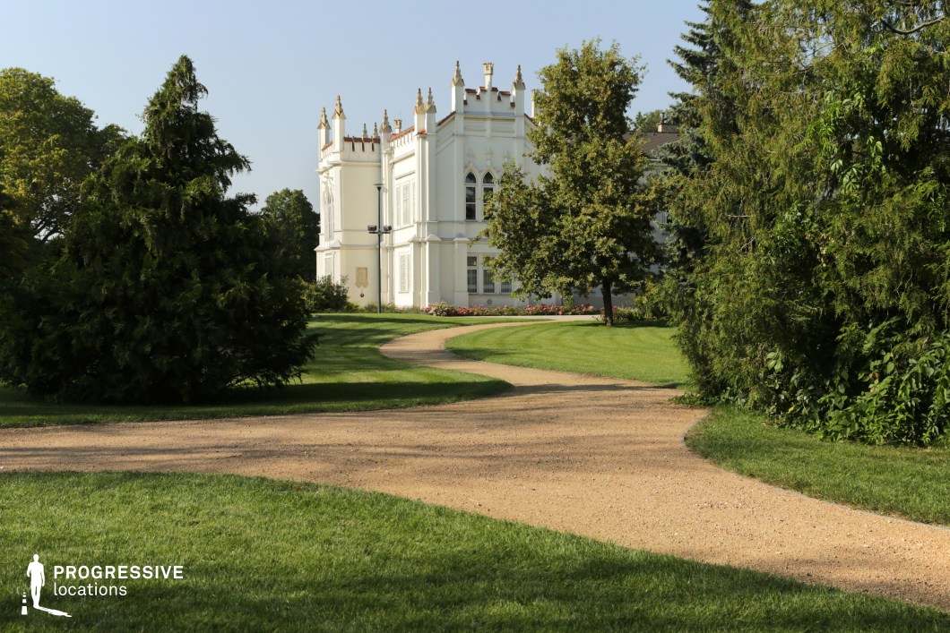 This image features a gracefully curving gravel pathway winding through a lush, manicured garden towards an elegant white Gothic Revival mansion. The building’s decorative crenellations, pointed windows, and ornate detailing enhance its fairytale-like character, while the surrounding greenery creates a secluded and tranquil atmosphere. The well-maintained lawn and carefully arranged plants add to the refined aesthetic. This location is ideal for filming romantic period dramas, fantasy films, or historical narratives, providing a dreamy and elegant setting with a strong visual impact.