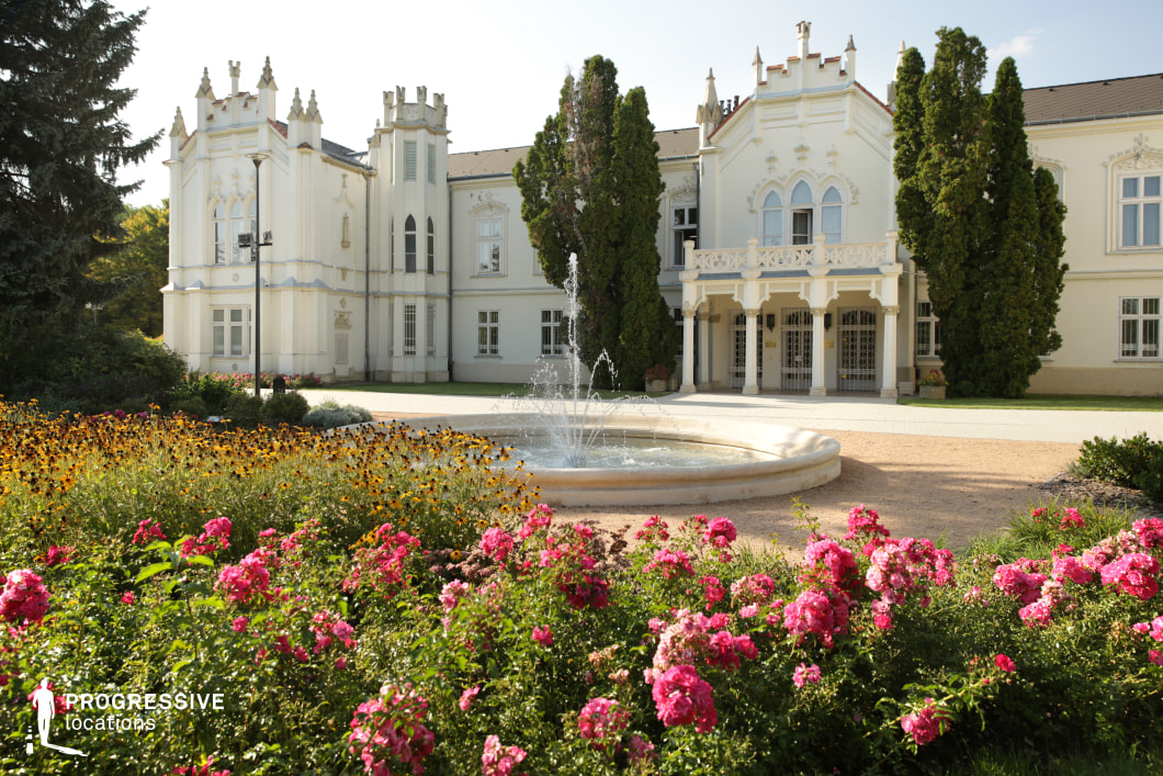 This image features a picturesque Gothic Revival-style mansion with pointed arches, decorative battlements, and an ornate balcony, exuding a fairytale-like charm. The pristine white facade is accentuated by intricate detailing, tall windows, and lush greenery. A central fountain with cascading water adds elegance to the courtyard, while the vibrant flower garden in the foreground enhances the scene’s romantic and idyllic atmosphere. This location is ideal for filming period dramas, fantasy films, or romantic storylines, offering a refined and enchanting setting for aristocratic gatherings, serene retreats, or timeless love stories.