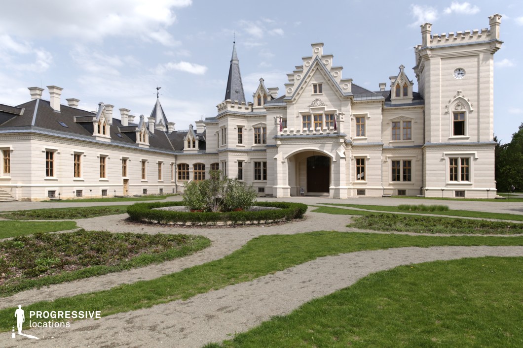 The image showcases a grand neo-Gothic style mansion with intricate architectural detailing, including pointed turrets, decorative gables, and arched windows. The building's light-colored stone façade contrasts with its dark, steeply pitched roof, creating a striking visual appeal. The symmetrical design is complemented by a central arched entranceway, flanked by a crenelated tower on one side, adding a medieval-inspired aesthetic. The surrounding landscaped garden features geometrically arranged greenery and winding gravel pathways, leading towards the main building. This location is ideal for filming period dramas, historical narratives, or fantasy-themed productions, offering a stately and timeless backdrop suitable for regal settings or mysterious, aristocratic storylines.