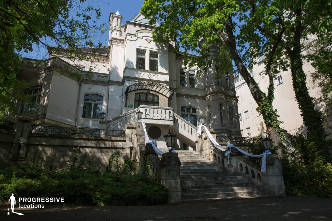 This image features a stately historic mansion with ornate architectural details, including decorative moldings, arched windows, and a turret-like structure that enhances its grandeur. The central stone staircase, flanked by elegant wrought-iron railings and traditional lanterns, creates a dramatic entrance leading up to the building’s main doors. Lush greenery surrounds the structure, complementing its aged stone and white façade. The combination of intricate detailing and a slightly weathered appearance gives the location a timeless character, making it ideal for filming period dramas, mystery thrillers, or romantic films, offering a regal and atmospheric setting with strong visual storytelling potential.