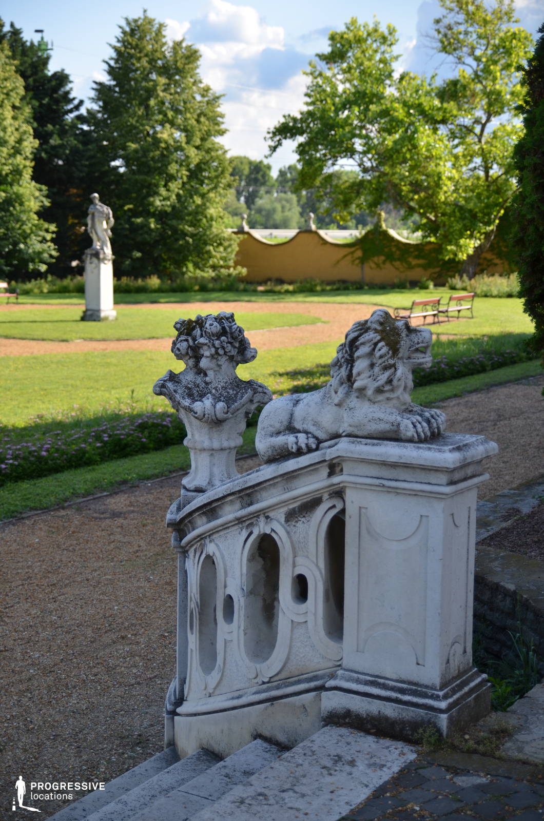 This elegant stone staircase features intricate carvings, including a reclining lion statue and a floral urn, adding a regal touch to the garden setting. The balustrade showcases delicate, arched cutouts that enhance the classical aesthetic. In the background, a manicured lawn and a gracefully curved garden wall with sculptural elements create a harmonious, historic atmosphere. A distant statue positioned along a gravel pathway adds depth and symmetry to the scene. This location is ideal for historical dramas, period films, and romantic settings, offering a refined and visually captivating backdrop for aristocratic or mysterious narratives.