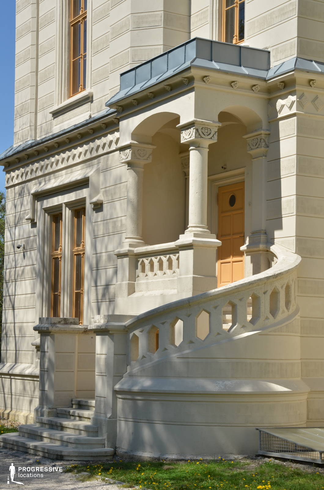 This refined architectural detail showcases a gracefully curved staircase leading to a small, columned entrance porch. The balustrade features a repeating geometric pattern, while the supporting columns and decorative friezes add a classical touch to the design. The pale stone facade is adorned with subtle ornamental reliefs, and the warm wooden door provides a contrasting element. Large windows with wooden frames enhance the sense of elegance and symmetry. This location is ideal for historical dramas, aristocratic settings, and period films, offering a sophisticated and visually engaging backdrop with a timeless European aesthetic.