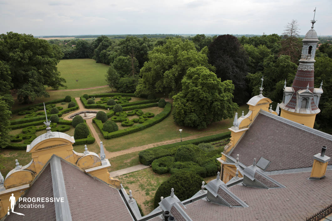 This image provides a high-angle perspective of a meticulously designed Baroque-style garden, framed by the ornate rooftops and spires of a historic palace. The symmetrical hedge formations and central fountain create a visually striking pattern, leading into a vast open lawn bordered by dense, mature trees. The rich architectural detailing of the palace roof, including decorative finials and intricate turrets, enhances the grandeur of the setting. This location is ideal for filming period dramas, aristocratic-themed productions, or romantic narratives, offering a refined and immersive historical backdrop with a sense of elegance and scale.