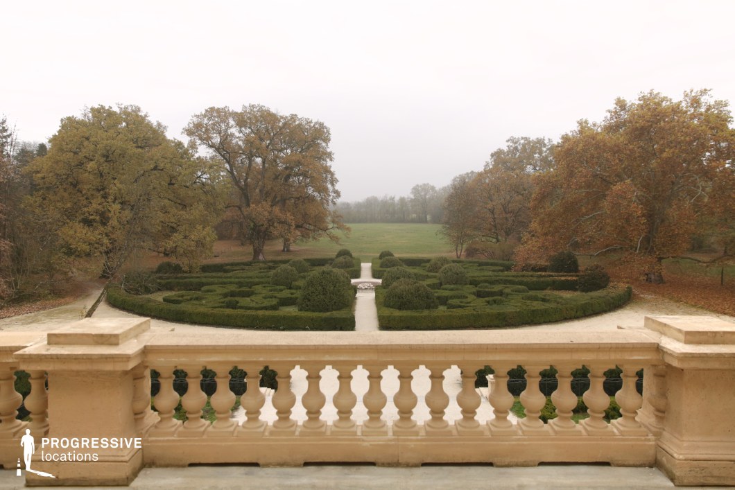 This image presents a meticulously designed formal garden viewed from an elevated terrace with a classic stone balustrade. The symmetrical hedge formations lead towards a central focal point, creating a structured and elegant visual composition. Beyond the garden, an open landscape stretches into the distance, framed by mature trees that add depth and contrast to the scene. The combination of the manicured greenery and the expansive natural surroundings makes this an ideal location for filming period dramas, romantic films, or historical narratives, offering a refined and atmospheric setting with a sense of timeless grandeur.