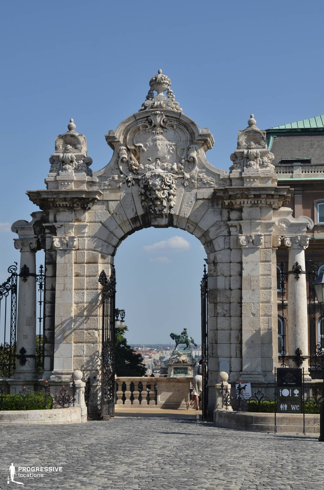 A grand baroque-style stone gate stands as the focal point, adorned with intricate carvings, sculpted reliefs, and decorative urns. The archway is flanked by elaborately wrought-iron gates, adding a sense of refinement and elegance. Beyond the arch, an equestrian statue is visible, positioned against a panoramic cityscape, creating a striking depth and perspective. The gate’s textured stone surfaces, combined with its classical architectural elements, exude historical grandeur. The cobblestone pathway leading up to the gate enhances the timeless aesthetic. This location is ideal for historical dramas, period films, and regal or aristocratic-themed productions, offering an authentic and visually captivating European backdrop.