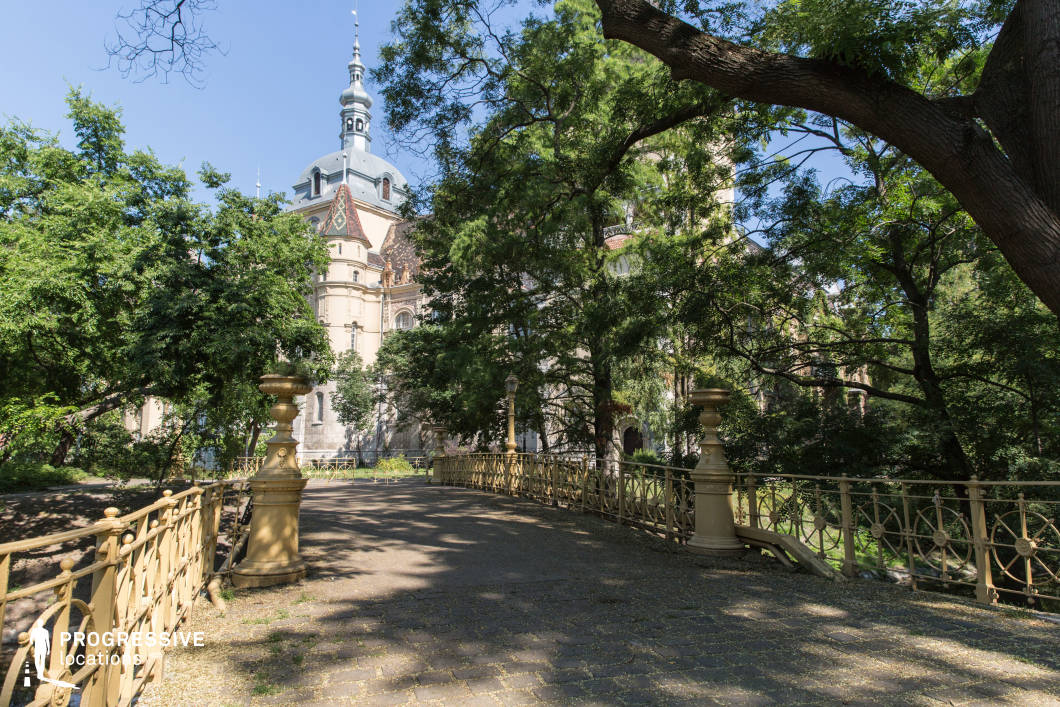 This image presents a striking historic castle with a mix of architectural styles, featuring a grand dome, intricate turrets, and steeply pitched roofs adorned with decorative tiles. The foreground showcases an elegant cast-iron bridge with ornamental railings and stone pillars, leading towards the castle, creating a visually engaging composition. Lush greenery surrounds the scene, partially obscuring the structure, adding an air of mystery and timeless charm. The combination of the castle's old-world European design and the picturesque bridge makes this location perfect for filming period dramas, fantasy adventures, or gothic-inspired narratives, offering a rich and immersive setting.
