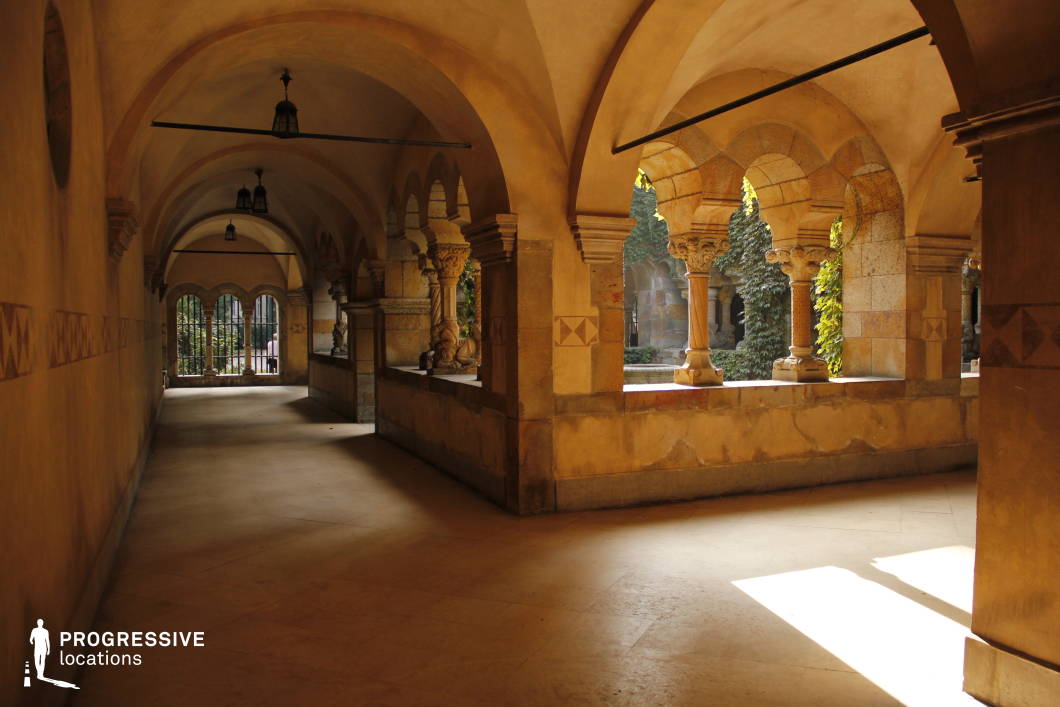 This image captures a beautifully preserved cloister with Romanesque and Gothic architectural elements. The covered walkway is lined with elegant stone columns featuring ornate capitals, supporting a series of rounded arches that create a rhythmic pattern of light and shadow. The warm, earthy tones of the stone walls and floor add to the aged, historical ambiance. The open archways provide glimpses of an inner courtyard, partially covered in creeping ivy, enhancing the secluded and timeless atmosphere. This location is ideal for filming medieval, historical, or fantasy-themed productions, offering a richly textured and immersive setting for mysterious, dramatic, or contemplative scenes.