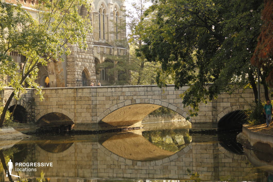 This image features an elegant stone bridge with arched openings, reflecting beautifully in the calm water below. The bridge’s textured masonry complements the imposing Gothic-style building in the background, which showcases pointed arches, intricate stonework, and medieval-inspired architectural elements. Lush greenery surrounds the scene, softening the solid stone structures and enhancing the tranquil yet dramatic atmosphere. The combination of the reflective water, aged stone surfaces, and historic architecture makes this location ideal for filming period dramas, fantasy narratives, or mystery films, offering a timeless and visually rich backdrop for atmospheric storytelling.